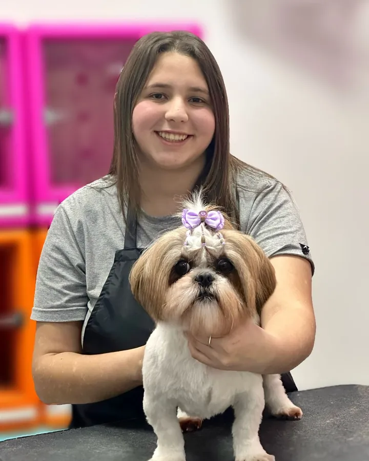 Aluna sorrindo atrás de um shih-tzu com laço lilás, de tosa pronta sobre a mesa