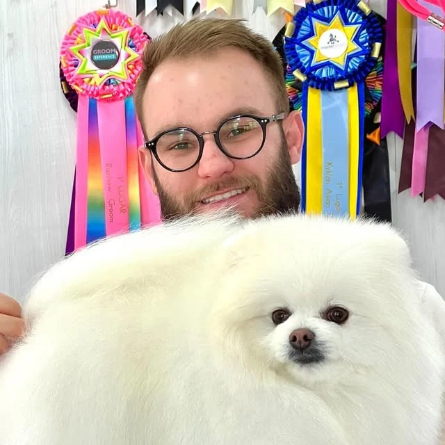 Gustavo Zilli sorrindo atrás de um spitz branco de pelagem cheia, com duas rosetas de primeiro lugar penduradas ao fundo